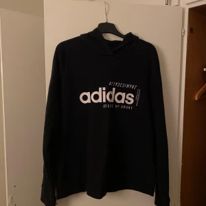 Svart hoodie från Adidas XL - Svart hoodie från Adidas i storlek XL med stor vit logga och text på bröstet. Tröjan har huva med snörning och långa ärmar. Mjuk och skön känsla, perfekt för chill eller träning. Klassisk och enkel design som funkar till många stilar.