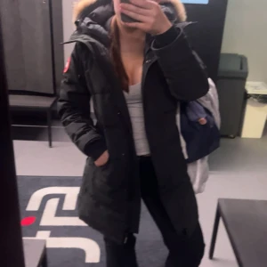 Svart lång dunjacka Canada Goose - Säljer en svart lång dunjacka från Canada Goose med ikonisk patch på ärmen och fluffig päls runt huvan. Jackan har dragkedja och knappar framtill, två fickor och justerbar huva. Perfekt för kalla vinterdagar och riktigt snygg streetstil.