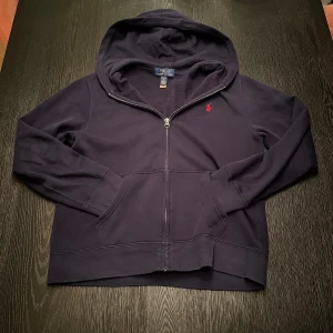 Mörkblå hoodie från Polo Ralph Lauren - Snygg mörkblå hoodie från Polo Ralph Lauren med dragkedja och huva. Tröjan har ett rött broderat Polo-logo på bröstet, två fickor fram och är tillverkad i mjuk bomull. Perfekt för chill dagar och enkel att matcha med jeans eller joggers.