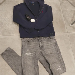 Mörkblå hoodie från Polo Ralph Lauren - Snygg mörkblå hoodie från Polo Ralph Lauren med röd broderad logga på bröstet. Tröjan har huva, dragkedja framtill och två praktiska fickor. Tillverkad i mjuk bomull, perfekt för chill dagar. Klassisk och stilren design som funkar året runt.