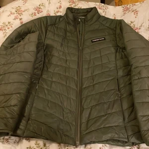 Grön pufferjacka från Cross Sportswear - Snygg mörkgrön pufferjacka från Cross Sportswear i storlek L. Jackan har quiltad design, hög krage och dragkedja framtill samt två sidofickor med dragkedja. Materialet är 100% nylon vilket gör den lätt och smidig. Perfekt för kyliga dagar.Mera som en M.