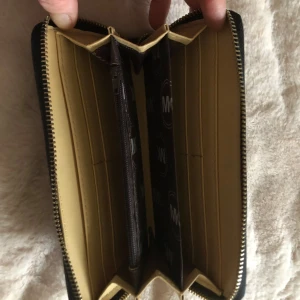 Svart plånbok från Michael Kors - Snygg svart plånbok från Michael Kors med guldfärgad logga på framsidan. Plånboken har dragkedja runtom och flera fack för kort och sedlar samt ett myntfack med zip. Utsidan är i stilrent skinn och insidan är beige med MK-mönster.