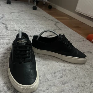 Svarta Saint Laurent sneakers i skinn - Svarta sneakers från Saint Laurent Paris med vit platt sula och klassisk snörning. Skorna är tillverkade i mjukt skinn och har en rund tå. Diskret logga på plösen och yttersulan. Perfekt för dig som gillar stilrena och lyxiga sneakers.