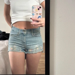 Levi's ljusblå jeansshorts med broderi - Säljer ett par ljusblå jeansshorts från Levi's med hög midja och råa, fransiga kanter. Shortsen har broderade blommor i rosa, vitt och gult framtill, klassiska fem fickor och Levi's patch bak. Perfekta för sommarens chill dagar!