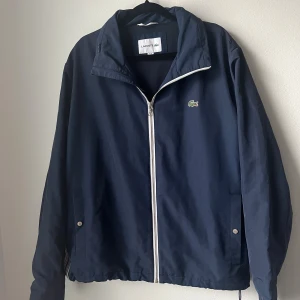 Marinblå vindjacka från Lacoste - Snygg marinblå vindjacka från Lacoste med klassisk krokodillogga på bröstet. Jackan har vit dragkedja, hög krage och två fickor med tryckknappar. Materialet är lätt och syntetiskt, perfekt för blåsiga dagar. Justerbar nederkant med snörning för extra passform. Som nytt skick! Storlek S