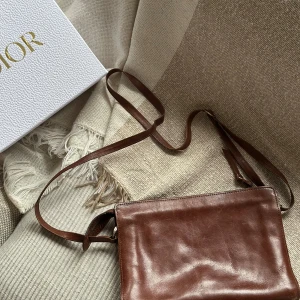 Brun axelväska i skinn med flätad detalj - Snygg brun axelväska i äkta skinn med lång rem och rektangulär form. Väskan har en flätad detalj på framsidan och flera fack med dragkedja inuti, perfekt för att hålla ordning på dina grejer. Stilren och klassisk design som funkar till många outfits.