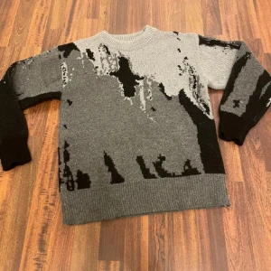 Grå och svart stickad tröja - Säljer en stickad tröja i grått och svart med abstrakt mönster. Tröjan har rund halsringning, ribbade muddar och lång ärm. Perfekt för dig som gillar en unik och cool stil med streetkänsla.
