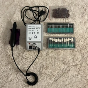 Elektrisk nagelfil med tillbehör - Komplett elektrisk nagelfil med kontrollenhet, handstycke, hållare, sliprullar och två set med olika filhuvuden. Perfekt för manikyr och pedikyr. Allt i gott skick, inga synliga defekter eller slitage. Enkel att använda och passar både hemma och professionellt.