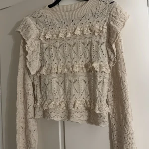Beige stickad tröja med volanger - Superfin beige stickad tröja med lång ärm och detaljerade volanger i spets längs bröstet och axlarna. Tröjan har ett snyggt hålmönster och ribbad rund halsringning. Sminkfläck vid halsen 