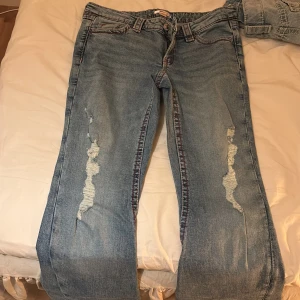 Blå slitna jeans från Gina Tricot - Ett par ljusblå jeans från Gina Tricot med slitna detaljer på båda benen och röda sömmar längs sidorna. Klassisk femficksmodell i bomull med rak passform och snyggt distressed look. Perfekta för en avslappnad och trendig stil.