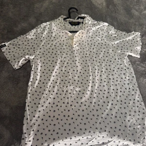 Vit blommig henley från Polo Golf - Vit henley-skjorta från Polo Golf Ralph Lauren med små svarta blommor över hela tyget. Skjortan har korta ärmar, knappar framtill och en klassisk krage. Tillverkad i mjuk bomull, perfekt för dig som gillar stilren design med en twist. Liten fläck på den med