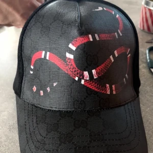 Svart Gucci keps med röd orm - Snygg svart keps från Gucci med diskret GG-mönster och en stor röd och vit orm på framsidan. Kepsen har klassisk form med böjd skärm och mesh på sidorna för extra ventilation. Perfekt för dig som vill sticka ut med designerstil.