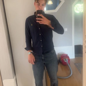 Mörkblå skjorta från Polo Ralph Lauren - Snygg mörkblå skjorta från Polo Ralph Lauren i slim fit med klassisk krage och knappar framtill. Ikonisk gul broderad logga på bröstet. Skjortan är långärmad och tillverkad i mjuk bomull, perfekt för en clean och stilren look.