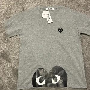 Grå Comme des Garçons Play t-shirt - Snygg grå t-shirt från Comme des Garçons Play med ikoniskt svart hjärta på bröstet och stor svart/vit hjärtdetalj längst ner. Klassisk passform och rund hals. Perfekt för dig som gillar streetstyle och vill sticka ut med en unik detalj. säljer pga att den var för liten för mig 
