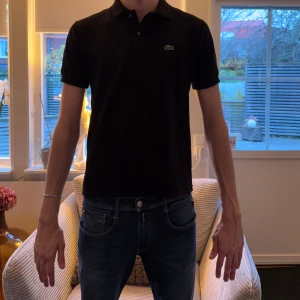 Svart piké från Lacoste classic fit - Svart pikétröja från Lacoste i klassisk passform. Tröjan har korta ärmar, krage med två knappar och den ikoniska krokodilloggan på bröstet. Materialet är mjuk bomull som känns skönt mot huden. Perfekt för dig som gillar stilrena och tidlösa plagg.