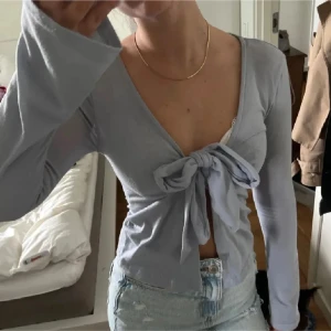 Ljusblå omlottopp med knyt framtill - Säljer en ljusblå omlottopp med lång ärm och stor knytrosett framtill. Toppen har v-ringad hals och är croppad, vilket ger en snygg och trendig look. Materialet känns mjukt och stretchigt, perfekt att styla med jeans eller kjol.