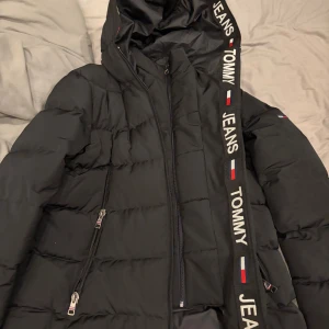 Svart pufferjacka Tommy Jeans M - Svart pufferjacka från Tommy Jeans med huva och dragkedjor framtill. Jackan har breda quiltade partier, två fickor med dragkedja och tydliga logodetaljer på huvan. Perfekt för kyliga dagar och riktigt snygg streetstil.