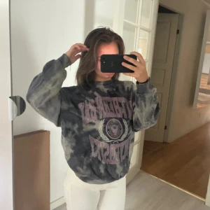 Grå tie-dye sweatshirt från Pull&Bear - Säljer en oversized grå och svart tie-dye sweatshirt från Pull&Bear med tryck i lila och vitt framtill. Tröjan har rund hals, ribbade muddar och lång ärm. Perfekt för dig som gillar streetstyle och comfy vibes.