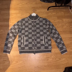 Grå stickad kofta från Gucci - Säljer en grå stickad kofta från Gucci med vit GG-logga över hela tröjan. Koftan har dragkedja framtill, PRIS GÅR ATT DISKUTERAS
