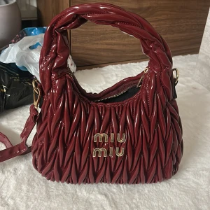 Vinröd axelväska från Miu Miu - Snygg vinröd axelväska från Miu Miu med glansigt, quiltat skinn och guldfärgade detaljer. Väskan har ett unikt tvinnat mönster, bred axelrem och dragkedja upptill. Perfekt storlek för det viktigaste och ger en lyxig vibe till din outfit.
