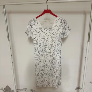 Vit kortklänning med spets och mönster - Vit kortklänning med svart prickigt mönster och rund halsringning. Klänningen har korta ärmar och en snygg spetsdetalj vid halsen som ger en extra touch. Materialet känns mjukt och lätt, perfekt för varmare dagar. Stilren och modern design som sticker ut.