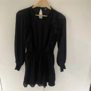 Svart volangklänning från Zara - Supersöt svart kortklänning från Zara med volanger längs sidorna och prickigt mönster i tyget. Klänningen har långärmade ärmar med smock vid handlederna, markerad midja och rund hals med spetsdetaljer. Nyckelhålsöppning bak i nacken. Perfekt för dig som gillar romantisk stil.