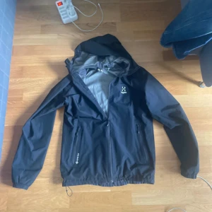 Svart vindjacka från Haglöfs - Säljer en svart vindjacka från Haglöfs med huva och dragkedja framtill. Jackan har två sidofickor med dragkedja och justerbar nederkant. Perfekt för blåsiga dagar och outdoor-aktiviteter. Materialet är slitstark polyester och jackan har en stilren look. Den är köpt för 3000kr, pris kan diskuteras 
