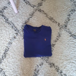 Blå t-shirt från Polo Ralph Lauren - Snygg blå t-shirt från Polo Ralph Lauren med klassisk orange logga broderad på bröstet. T-shirten har rund halsringning och korta ärmar. Tillverkad i mjuk bomull som känns skön mot huden. Perfekt för en clean och stilren look. Strl: passar någon som är 155. 