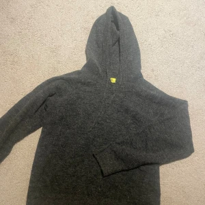 Mörkgrå stickad hoodie med huva - Mysig mörkgrå stickad hoodie med huva och långa ärmar. Perfekt för kyliga dagar och enkel att matcha med jeans eller mjukisbyxor. Klassisk modell utan tryck eller detaljer, bara ren och enkel stil.