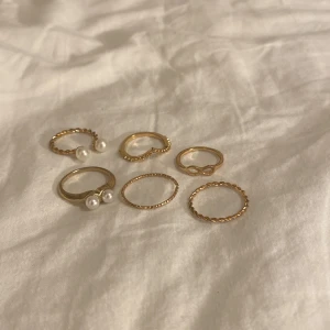 Set med sex guldiga ringar med pärlor - Säljer ett set med sex tunna guldiga ringar i olika former, vissa med tvinnad design och två med vita pärldetaljer. En ring har hjärtformad kant och en annan har en infinity-symbol. Perfekt för att mixa och matcha på fingrarna.