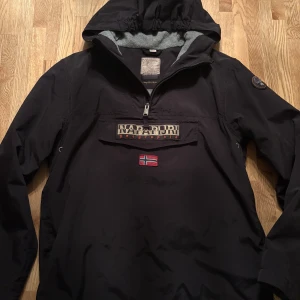 Svart vindjacka från Napapijri - Svart vindjacka från Napapijri med stor logotyp och norsk flagga på bröstet. Jackan har huva, halv dragkedja och patch på ärmen. Passar 162-168 beroende på kroppsform. Köpt för 1800kr