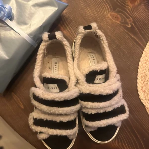 Svarta sneakers från Inuikii med fuskpäls - Unika svarta sneakers från Inuikii med fluffiga vita detaljer i fuskpäls och breda kardborreband. Skorna har en platt vit sula och rund tå, vilket ger dem en cool och mysig vibe. Perfekta för dig som gillar statement-skor med extra komfort och stil. Kartong finns med och de är använda kanske 5 gånger 