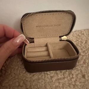 Brun smyckesask från Muli Collection - Snygg och kompakt smyckesask från Muli Collection i mörkbrunt skinn med guldfärgad dragkedja. Perfekt för att förvara ringar och små smycken. Insidan är beige och har fack för olika smycken. Stilren design som är lätt att ta med sig.