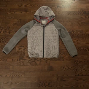 Grå pufferjacka från Jack & Jones - Snygg grå pufferjacka från Jack & Jones med huva och svarta detaljer. Jackan har quiltad framsida, mjuka grå ärmar och röd kant på insidan. Perfekt för kyliga dagar och har dragkedja framtill samt två fickor.