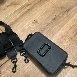 Svart axelväska från Marc Jacobs - Snygg, svart axelväska från Marc Jacobs med bred justerbar axelrem med logga. Väskan är rektangulär, har dubbla dragkedjor. Framsidan pryds av Marc Jacobs ikoniska logga i svart. 