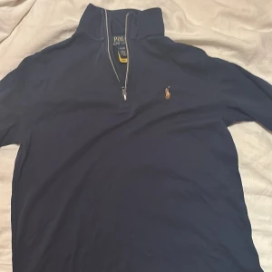 Marinblå half zip tröja Polo Ralph Lauren - Säljer en marinblå långärmad tröja från Polo Ralph Lauren med klassisk broderad logga på bröstet. Tröjan har en hög krage och half zip-dragkedja framtill. Tillverkad i mjuk bomull, perfekt för lager-på-lager under höst och vinter. Ganska liten i storlek. 