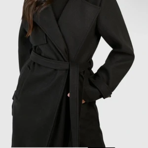 Svart lång kappa med bälte - Säljer en svart, lång kappa med breda slag och knytbälte i midjan. Jackan har långa ärmar med snygga knappdetaljer vid mudden och en stilren, omlott design. Perfekt för dig som gillar minimalistisk och clean stil.