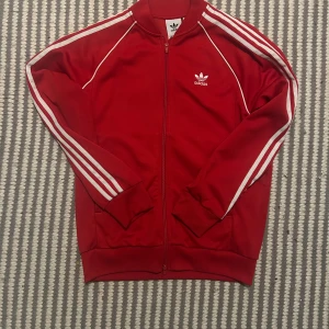 Röd Adidas vindjacka med vita ränder - Säljer en röd Adidas vindjacka med klassiska vita ränder längs ärmarna och broderad logga på bröstet. Jackan har dragkedja framtill, ribbade muddar och sportig look. Perfekt för dig som gillar streetwear och retrostil.