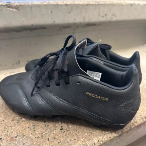 Svarta Adidas Predator fotbollsskor - Säljer ett par svarta Adidas Predator fotbollsskor med snygga guldfärgade detaljer och snörning. Skorna har en rund tå och är tillverkade i syntetmaterial med diskret perforerat mönster på sidorna. Perfekta för dig som vill ha stil på planen.