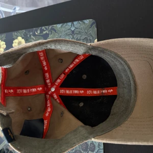 Beige keps Alpha Industries - Snygg beige keps från Alpha Industries med röd logotyp framtill på ett grönt patch. Justerbar baktill och tillverkad i slitstark bomull. Perfekt för dig som vill ha en clean och street-inspirerad look.