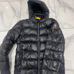 Svart dunjacka Parajumpers Sheen S - Svart dunjacka från Parajumpers, modell Sheen, med huva och coolt gult spänne vid halsen. Litet hål som knappt syns annars helt ny. Jackan har quiltad design, dragkedja framtill och patch på ärmen. Perfekt för kalla dagar och har en snygg, sportig look.