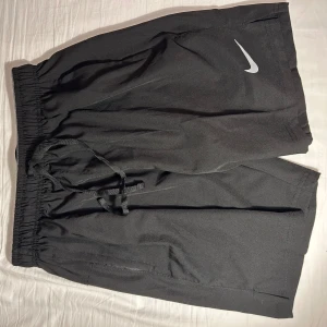 Svarta Nike Dri-FIT shorts M - Svarta sportiga shorts från Nike med Dri-FIT-teknologi. De har elastisk midja med snörning och diskret Nike-logga framtill. Perfekta för träning eller chill, tillverkade i lätt och snabbtorkande syntetmaterial.