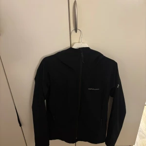 Svart vindjacka Peak Performance - Svart vindjacka från Peak Performance med huva och hel dragkedja framtill. Jackan har stilren design, diskret logga på bröstet och är tillverkad i syntetmaterial. Perfekt för dig som gillar sportig och clean look.