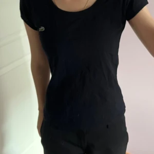 Svart kortärmad topp från Lacoste - Svart topp från Lacoste med rund halsringning och korta ärmar. Diskret Lacoste-logga på bröstet och broderad text på ärmen. Tunn och mjuk bomull som sitter snyggt och har en normal passform. Perfekt för en clean och sportig look.