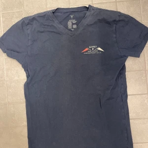 Mörkblå v-ringad t-shirt från GANT - Snygg mörkblå t-shirt från GANT med v-ringning och tryck på bröstet i vitt och rött. Klassisk passform och mjukt bomullsmaterial som känns skönt mot huden. Perfekt för dig som gillar stilrena och sportiga plagg med en touch av USA-vibe.