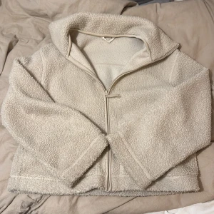 Beige teddyjacka med dragkedja - Mysig beige teddyjacka med hög krage och hel dragkedja framtill. Jackan har en fluffig och mjuk yta i fleece, perfekt för kyliga dagar. Enkel och stilren design som passar till många olika outfits.