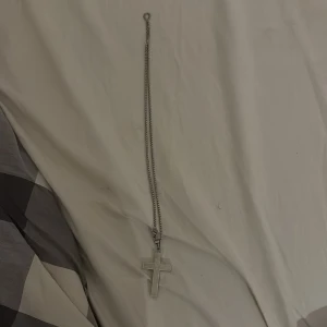 Silverfärgat halsband med korshänge - Stilrent halsband i silverfärg med ett korsformat hänge. Hänget har ingraverad text på framsidan och kedjan är av boxmodell med karbinlås. Perfekt för dig som gillar klassiska och tidlösa smycken med en personlig touch.