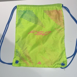 Neongul Adidas F50 påse - Snygg neongul gympåse från Adidas med F50-tryck och blå snören. Väskan har tunna ränder i blått och rosa samt dragskostängning. Perfekt för att bära träningskläder eller skor till gymmet eller fotbollen.