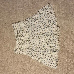 Vit blommig volangkjol - Supersöt kort kjol från 157 i vitt med små blå blommor och volangkant. Kjolen har smock i midjan för en bekväm passform och är perfekt för varma dagar. Tunt och lätt material som ger en luftig känsla.