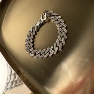 Glittrigt silverfärgat armband - Säljer ett chunky armband i silverfärg med glittrande stenar på varje länk. Armbandet har en bred kedjedesign som ger en lyxig och trendig känsla. Perfekt accessoar för att lyfta din outfit med extra bling. Köpt för 799